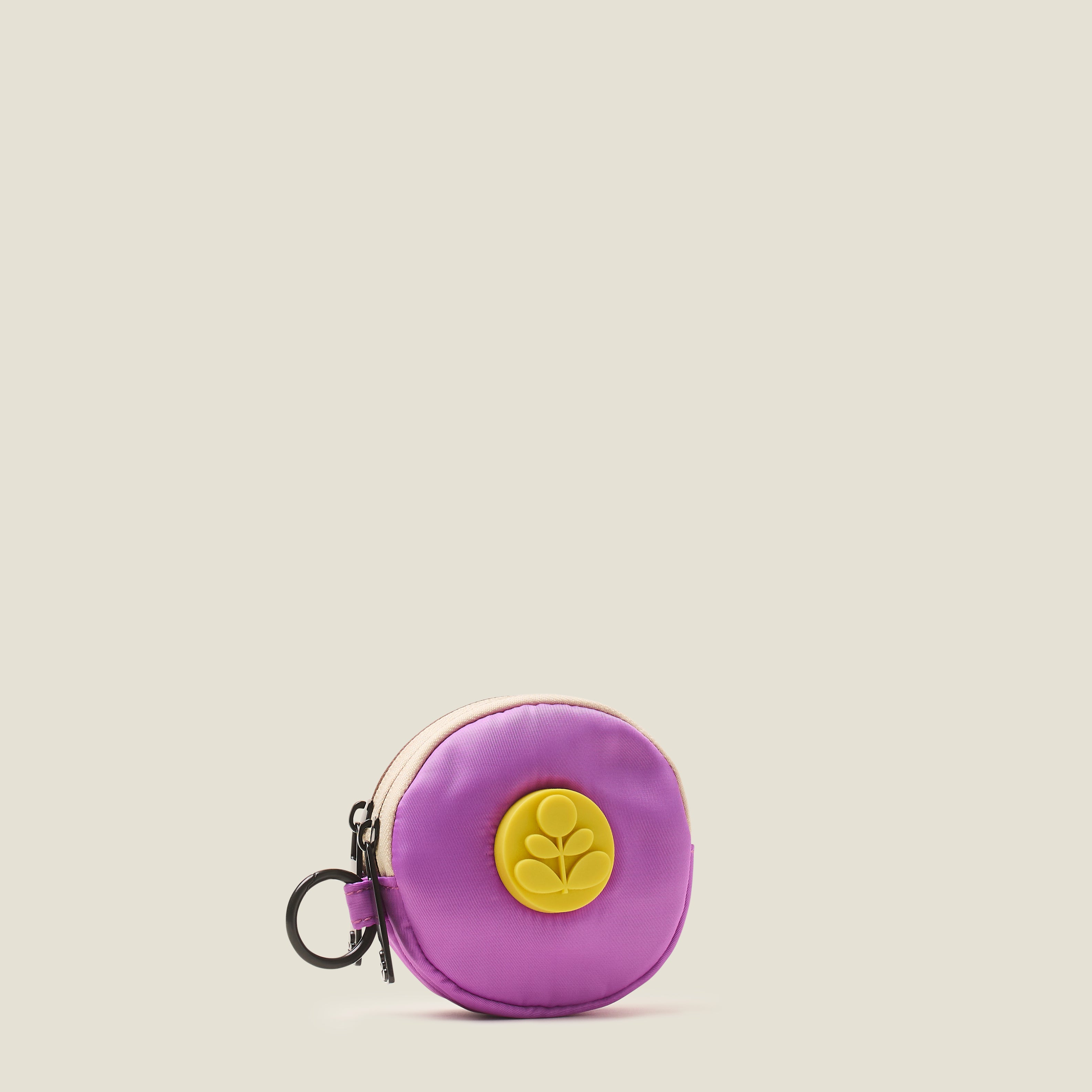 Side view of a lilac purple round coin purse with a rubber yellow Orla Kiely stem logo and ring clip on a beige background