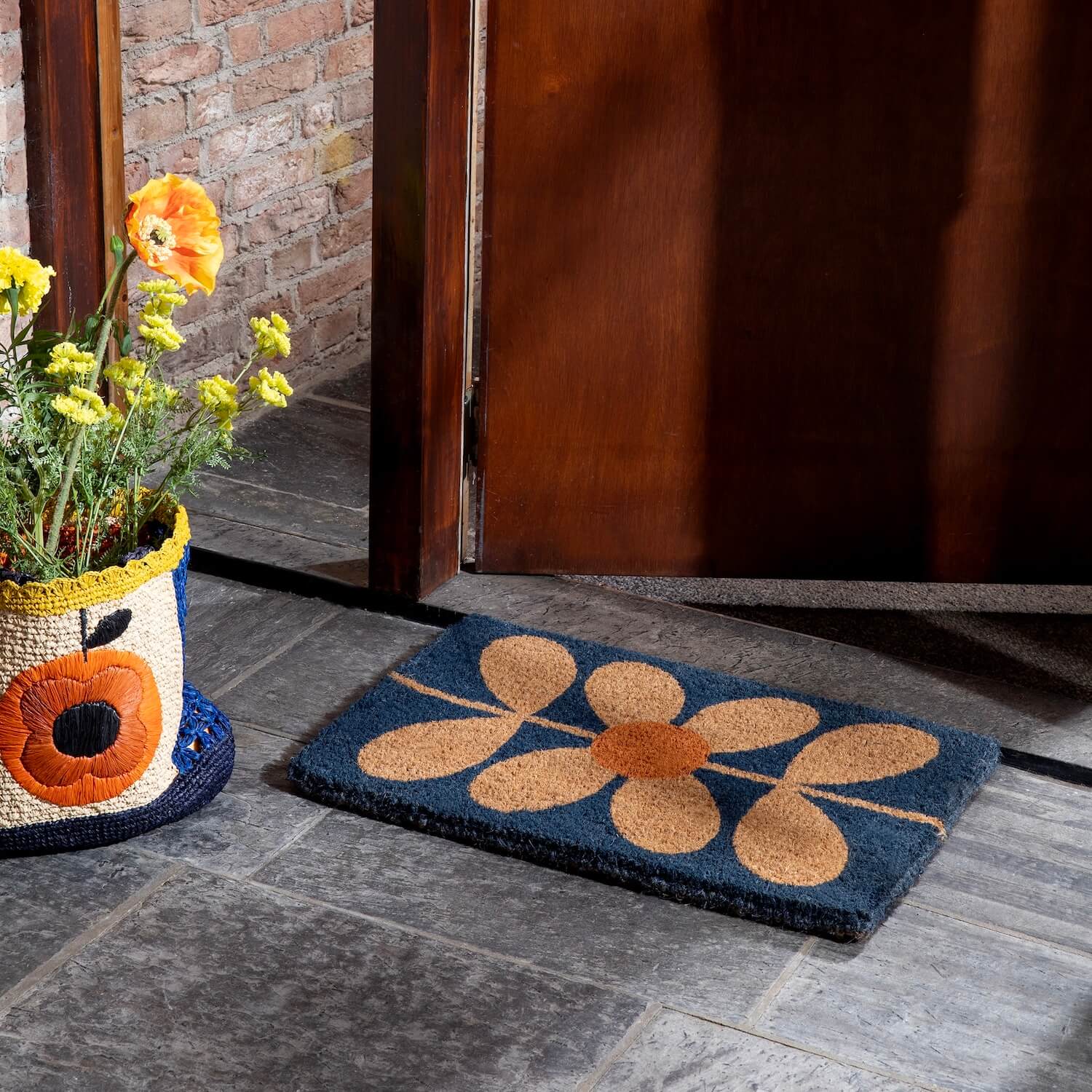 Lifestyle shot of Orla Kiely's sixties stem navy doormat next to a raffia bag